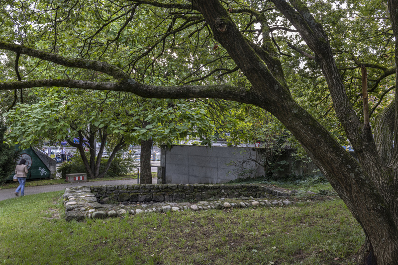 Der Blick geht von der Stadtmauer zum Rhein, im Vordergrund das wohl 1273 niedergebrannte Haus vor der Äußeren Stadtmauer. Die Bedeutung der Stadtmauern vor allem in fehdeartigen Kriegen – einer ernsthaften Belagerung hätten die Basler Stadtmauern wohl nicht standgehalten – zeigt sich an der Situation der Vorstädte. Vor dem Bau der äußeren Stadtmauer waren die Vorstädte Angriffen fast schutzlos ausgesetzt. Deshalb entstanden ab den 1370er-Jahren auch erste einfache Vorstadtbefestigungen. Rudolf von Habsburg überfiel 1253 das Kloster Maria Magdalena in der Steinenvorstadt und 1273 die St. Johanns-Vorstadt. Davon zeugt die kleine Hausruine mit Fundmaterial aus dem fraglichen Zeitraum (Pertz 1861, S. 195. Aebi; d’Aujourd’hui; Etter 1991).